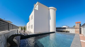En swimmingpool med en stenbelagd terrass, en vit byggnad med ett rundat hörn och en klarblå himmel.