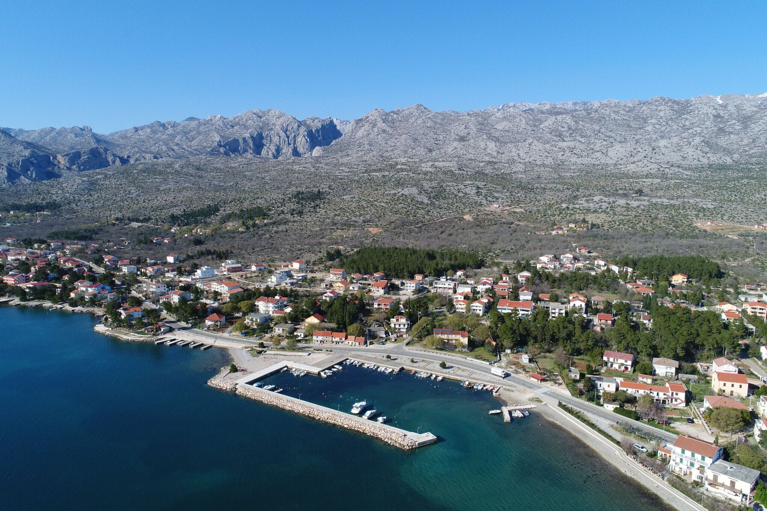 Vista aérea de un pueblo costero con un puerto deportivo, casas y montañas al fondo.