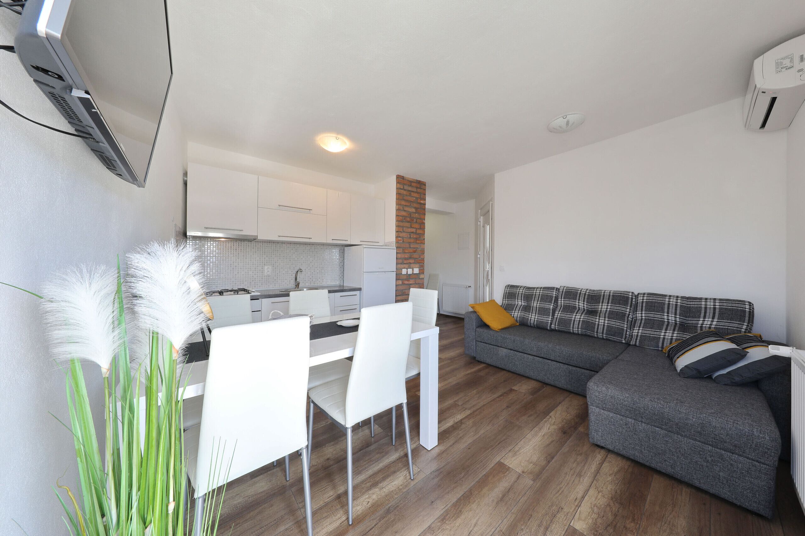 A modern living room with a grey sofa, white dining table, and chairs, a kitchen in the background, and a brick column.