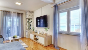 A modern living room with a flat-screen TV, wooden flooring, a stone accent wall, and a large window with curtains. - Apartment Pavao with Whirlpool (Rovanjska)