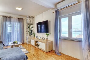 Ein modernes Wohnzimmer mit Flachbildfernseher, Holzboden, einer Wandverkleidung aus Stein und einem großen Fenster mit Vorhängen.