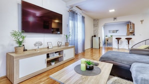 A modern living room with a flat-screen TV, a wooden console table, a grey sofa, and a dining area with bar stools. - Apartment Pavao with Whirlpool (Rovanjska)