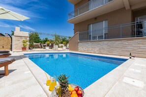 Una piscina con lettini, un cesto di frutta e un edificio con balconi.