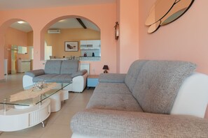 Ein Wohnzimmer mit grauem Sofa, einem Glastisch und einem weißen Beistelltisch. Im Hintergrund ist eine Küche mit weißen Schränken und einer Arbeitsplatte zu sehen.