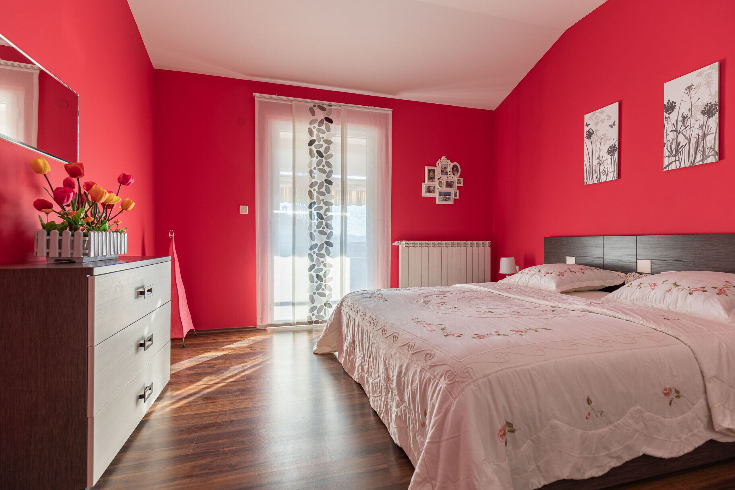 Ein Schlafzimmer mit roter Wand, einer Holzkommode, einem Bett mit Blumenmuster-Bettwäsche und einem Fenster mit dekorativer Vorhang.