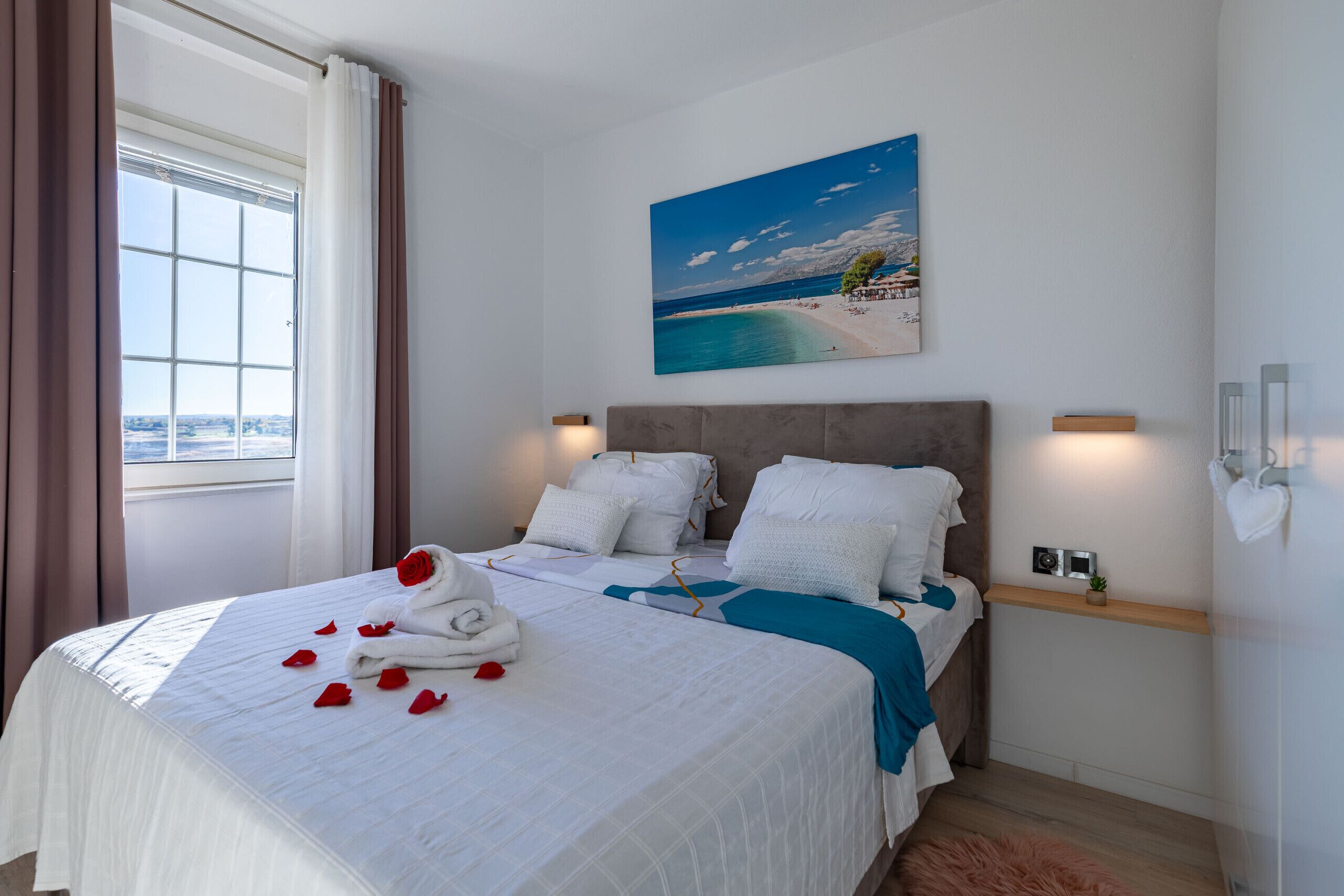 A hotel room with a bed, a painting of a beach, a window with curtains, and a nightstand with a lamp.