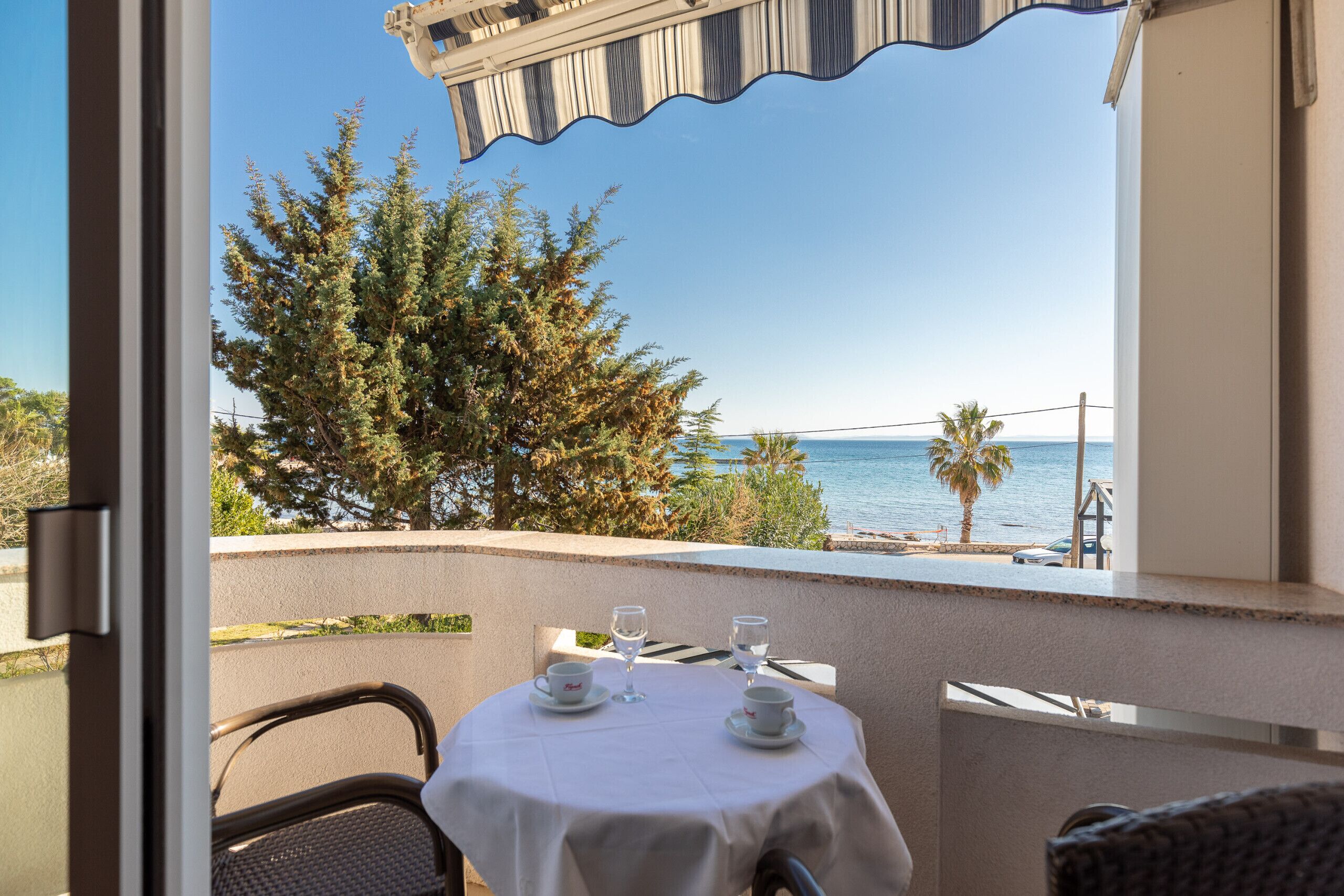 Un balcon avec une table dressée pour deux personnes, offrant une vue sur la côte, avec des arbres et un palmier.