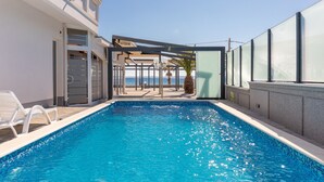 Una piscina con agua azul cristalina, un edificio blanco con balcón y vista al mar.
