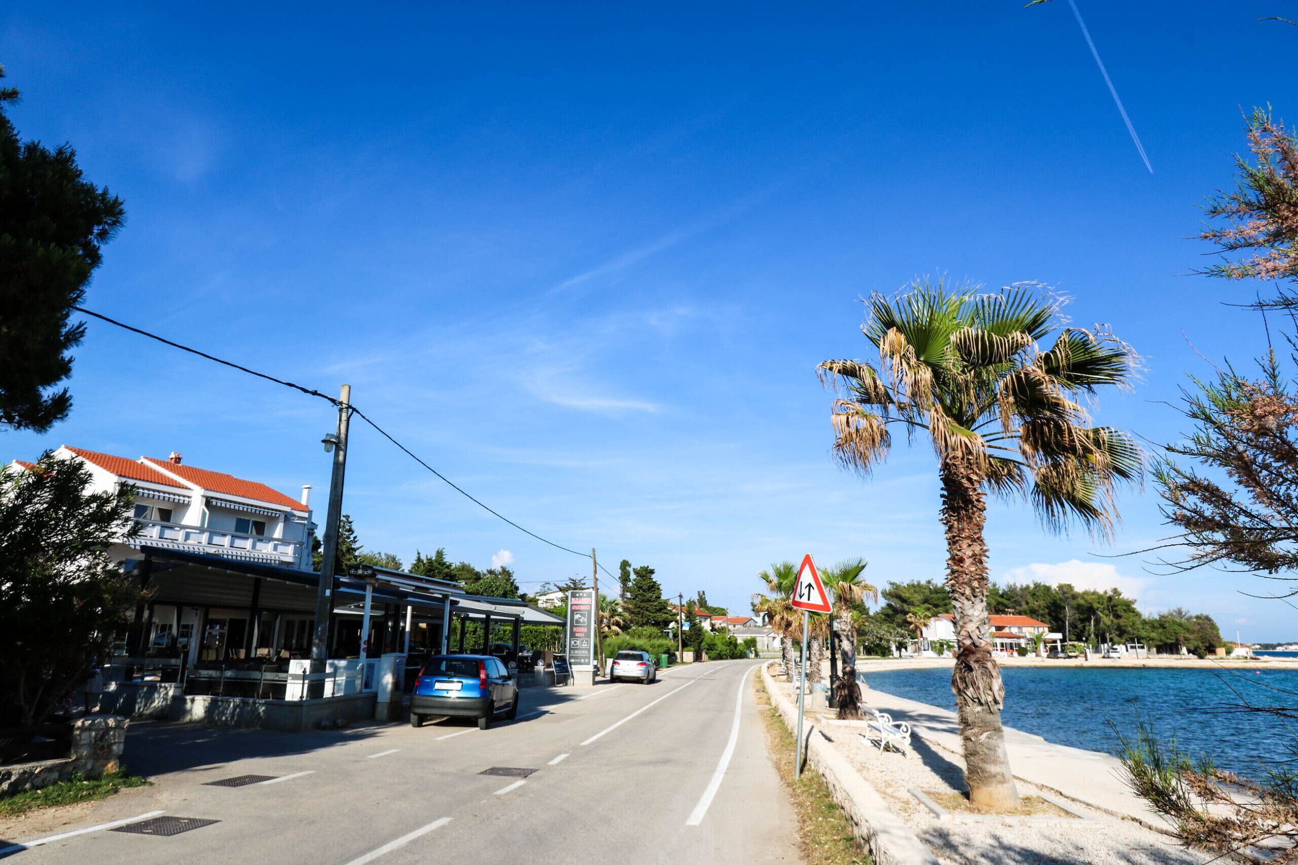 Une route côtière, avec un restaurant sur la gauche, un palmier sur la droite et une étendue d’eau en arrière-plan.