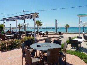 Ein Außenbereich mit Tischen und Stühlen, ein überdachter Bereich und Blick aufs Meer.