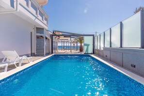 Ein Swimmingpool mit klarem, blauem Wasser, weißen Liegestühlen und Blick auf das Meer.
