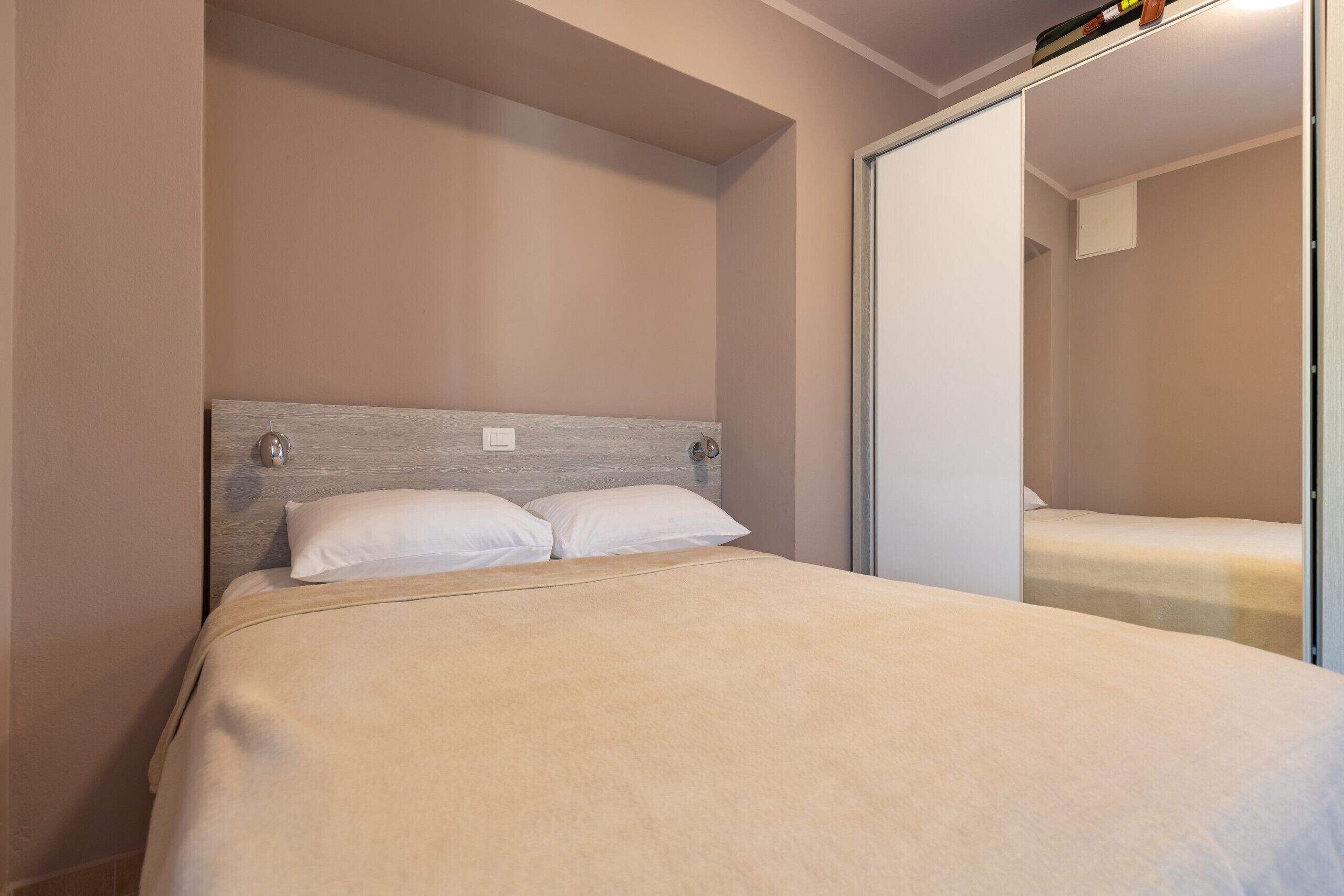 A single bed with a wooden headboard, a white pillow, and a neutral-colored bedspread in a room with a sliding door and a light switch on the wall.