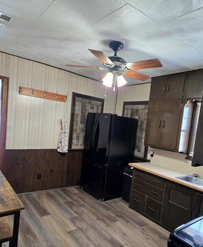 A kitchen with wooden cabinets, a black refrigerator, a ceiling fan, and a wooden table.