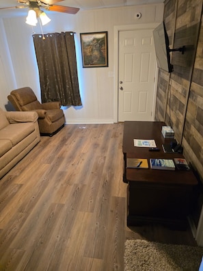 A room with a ceiling fan, a door, a framed picture, a brown armchair, a sofa, a wooden table with books, and a rug.