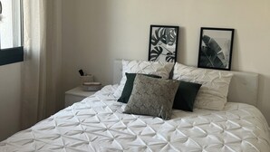 Una cama bien hecha con una colcha de estampado geométrico, una mesita de noche con lámpara y dos impresiones botánicas enmarcadas en la pared.