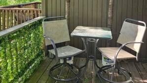 Eine Holzterrasse mit zwei Stühlen und einem Tisch, umgeben von einem Holenzaun und einem grünen Heckenstreifen.