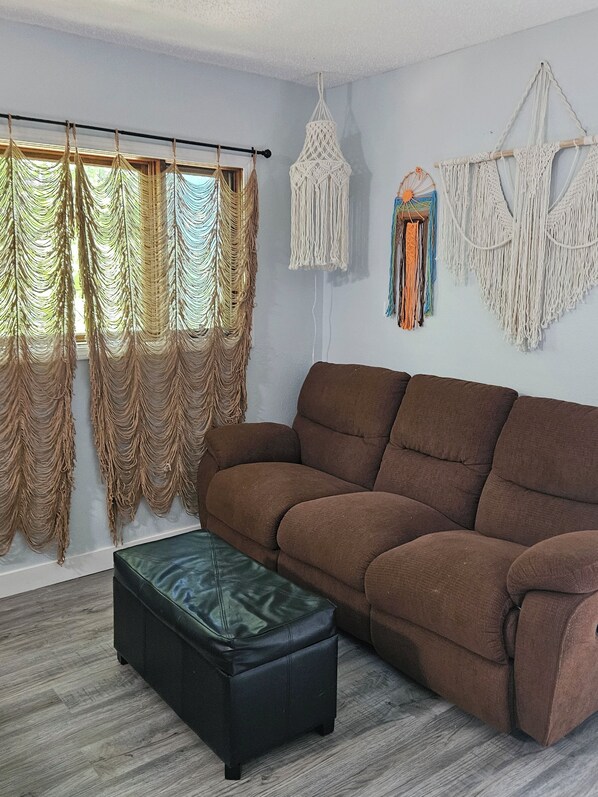 A living room with a brown sofa, a black ottoman, macramé wall hangings, and a window with curtains. - Welcome to the HIPPY HAVEN nothing but good vibes (Lake Ozark)