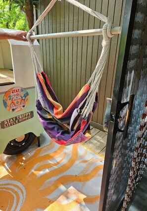 Una hamaca colgada de un poste metálico, un tapete colorido en el suelo y un vehículo con un adhesivo que dice 'Stay Trippy Little Hippie'.