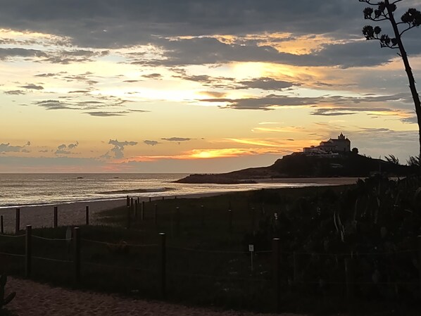 Une vue sur le coucher de soleil le long de la cÎte, avec une plage, une clÎture et un bùtiment situé sur une colline.