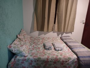 Un lit simple avec une literie Ă motifs floraux, une table de chevet sur laquelle est pliĂ©e une couverture, et une fenĂȘtre avec des rideaux beiges.