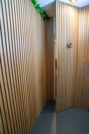 Un couloir avec des lambris en bois verticaux et un plancher foncé, qui mène à une porte portant l’inscription « WC ».