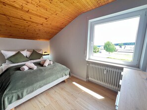 Une chambre avec un plafond en bois, une grande fenêtre, un lit avec des oreillers et un radiateur.