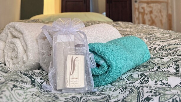 A bed with a patterned bedspread, a rolled white towel, a teal blanket, and a plastic bag with soap. - House in Progress (Colle di Val d'Elsa)