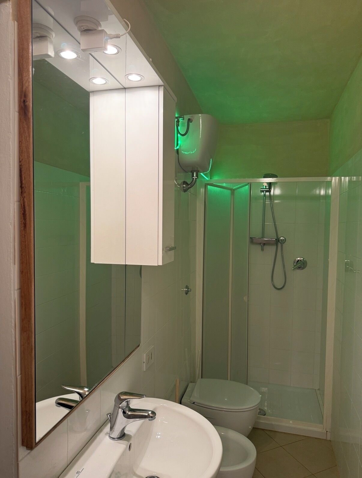 Een badkamer met een wit wastafel, spiegel en toilet, een douchecabine en een luchtzuiveraar aan het plafond.