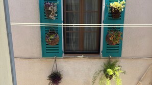 Uma varanda com vasos suspensos de flores, uma janela com persianas de madeira e vista para a rua.