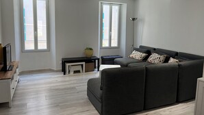 Uma sala de estar moderna com um sofá, uma mesinha e uma TV. Duas janelas proporcionam iluminação natural.
