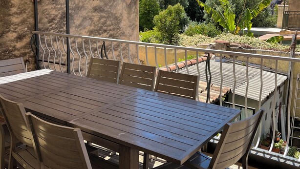 Um pátio externo com mesa e cadeiras, área coberta e vista para um jardim e edifícios.