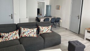 Uma sala moderna com um sofá cinzento, uma mesa de jantar com cadeiras e uma cozinha ao fundo.