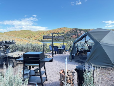 A camping setup with a tent, grill, chairs, and a table.. Unplug and unwind in our secluded off-grid glamping dome, nestled by a private lake. Enjoy the peace and quiet with all the comforts of home