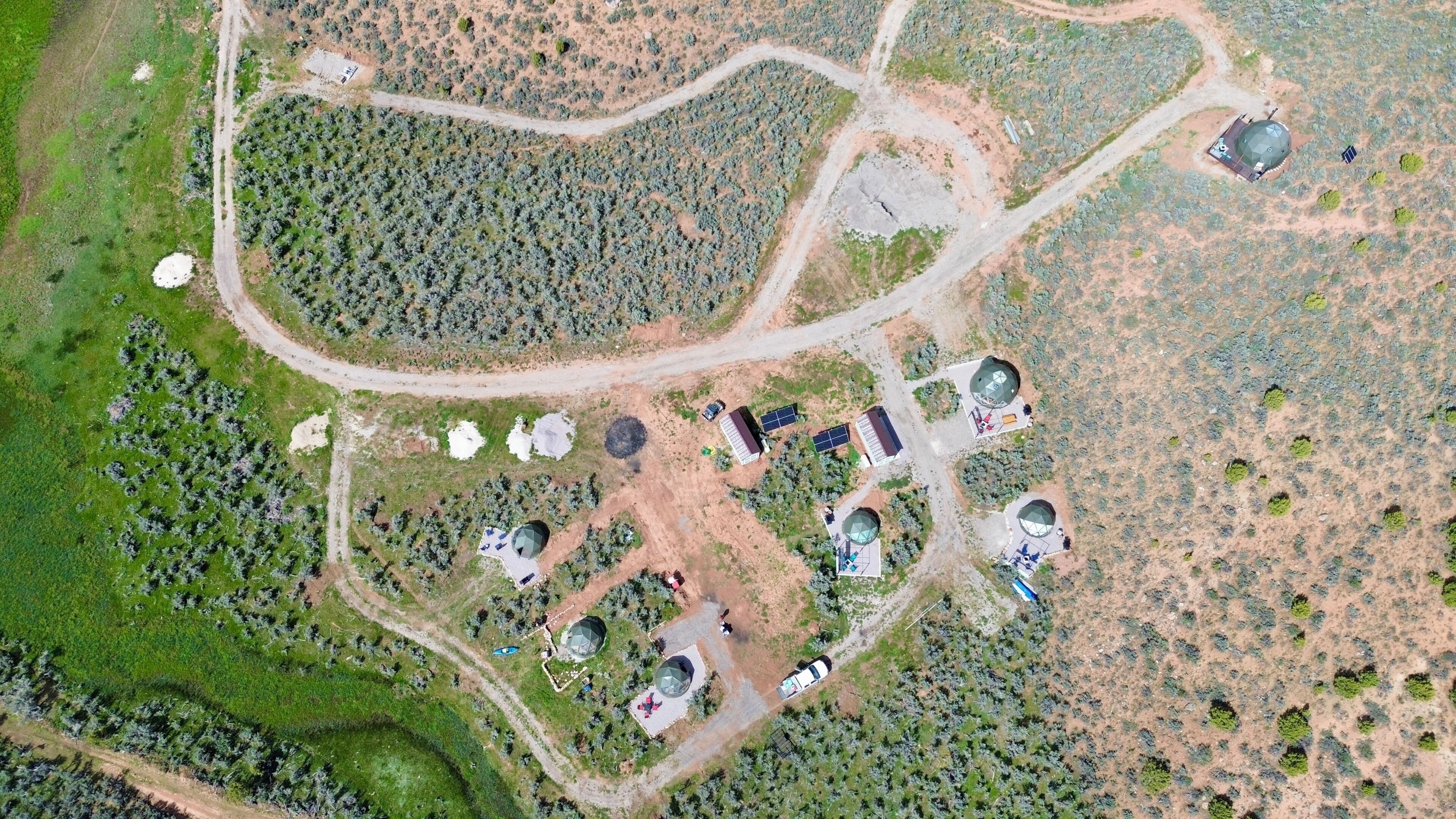An aerial view of a facility with multiple structures, including domed buildings and rectangular buildings, surrounded by sparse vegetation and a winding road.