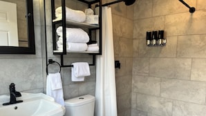 Ein Badezimmer mit Waschbecken, Toilette und Duschvorhang. Regale mit Handtüchern und Pflegeprodukten sind vorhanden.
