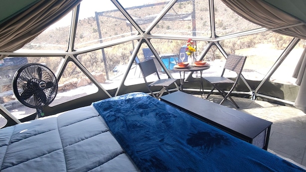 Una stanza con un letto, una ventola, un tavolo con sedie e vista su un paesaggio montuoso attraverso ampie finestre.