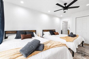 Quarto de hotel com duas camas, um ventilador de teto, paredes brancas e piso de madeira.