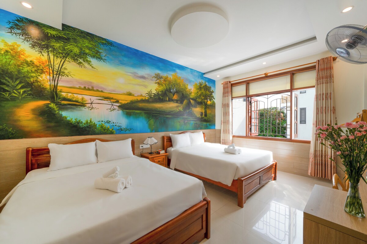 A hotel room with two beds, a large mural of a landscape, a window with curtains, and a desk with a vase of flowers.