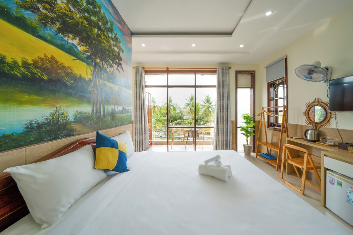 A hotel room with a large bed, a desk with a chair, a TV, and a view of greenery outside.
