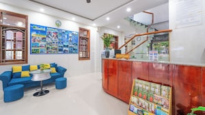 A reception area with a blue sofa, a glass-top table, a wooden reception desk, a clock, and a staircase. - New Moon Villa (Hoi An)