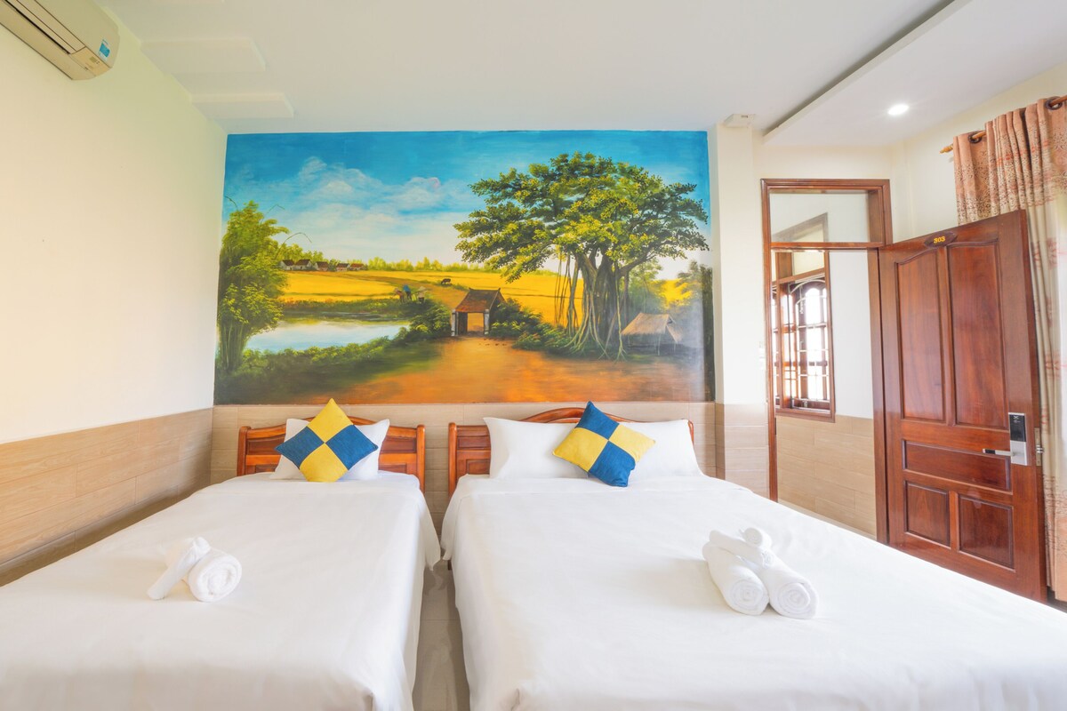 A hotel room with two beds, a painting of a landscape on the wall, a wooden door, and a window with curtains.