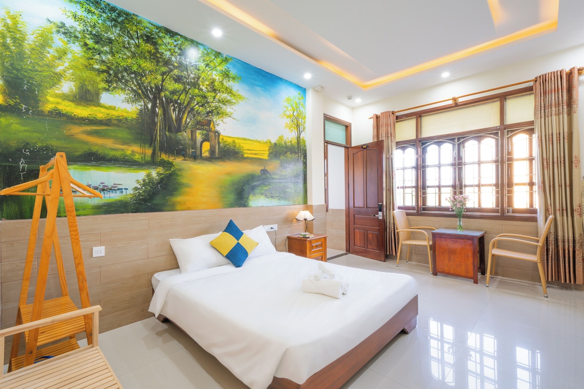A hotel room with a bed, a desk, a chair, a lamp, and a painting on the wall.