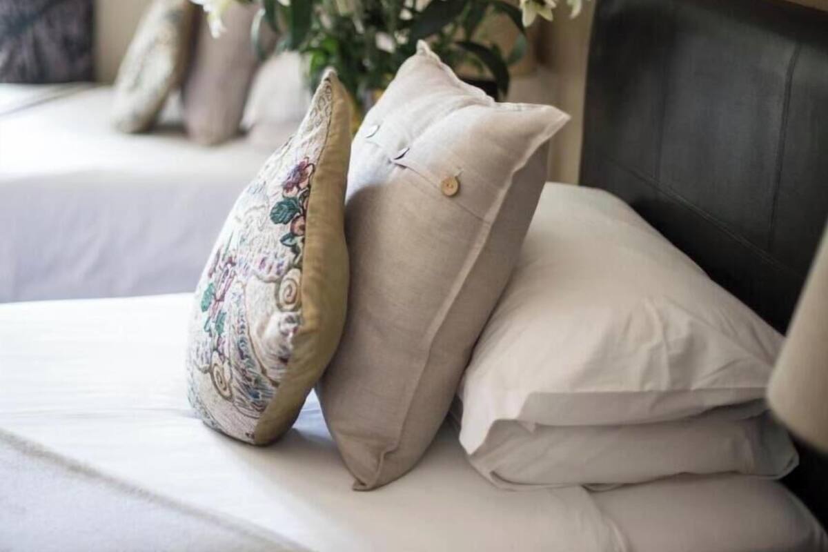 Un lit avec deux oreillers, l’un à motifs floraux et l’autre uni, dans une chambre dont la tête de lit est de couleur sombre, avec un vase de fleurs en arrière-plan.