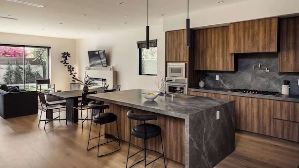 A modern kitchen with a large island, bar stools, wooden cabinets, and a view of the outdoors. - Tilden House | Designer Pool Home in West LA (Los Angeles)