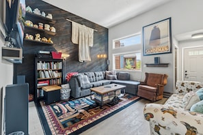 Ein modernes Wohnzimmer mit einer Eckcouch, einem Couchtisch, einem Bücherregal, einem gemusterten Teppich und einer Wandablage mit Dekoration.