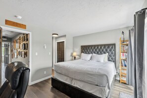 Ein Schlafzimmer mit einem großen Bett, einem Kopfteil, einem Nachttisch mit Lampe, einem Bücherregal und einer Tür zu einem anderen Raum.