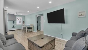 A modern living room with a TV, grey sofas, a wooden coffee table, and a kitchen area with a dining table and chairs. - Paxley`s: 2BR Spacious Beach Haven (Seaside Heights)