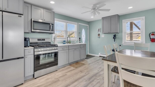 A modern kitchen with grey cabinets, a built-in microwave, stove, and oven. There is a dining table with chairs, a ceiling fan, and a window with blinds. - Paxley`s: 2BR Spacious Beach Haven (Seaside Heights)