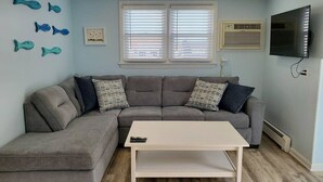 Ett vardagsrum med en grå hörnsoffa, ett vitt soffbord, en platt-TV och dekorativa fiskmotiv på väggen.