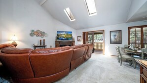 Un soggiorno con un divano in pelle marrone, una televisione e un camino. Sono presenti lucernari, una zona pranzo e vista sull'esterno.
